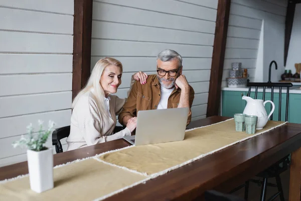 Elderly couple using laptop near jug and glasses on kitchen table — Stock Photo