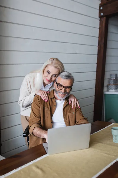 Seniorin umarmt lächelnden Ehemann in Brille mit Laptop neben Glas in Küche — Stockfoto