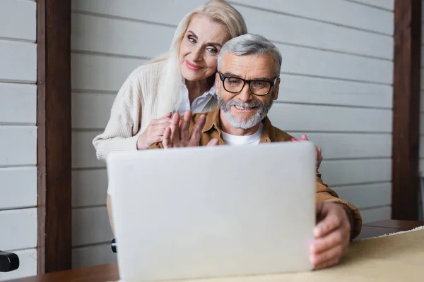 Elderly woman hugging husband waving hand during video chat on blurred laptop — Stock Photo