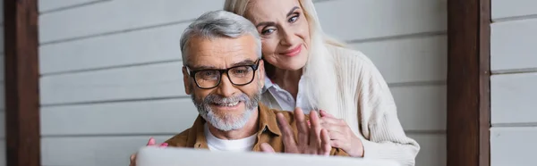 Улыбающийся пожилой мужчина машет рукой во время видеозвонка на размытом ноутбуке рядом с женой, баннер — стоковое фото