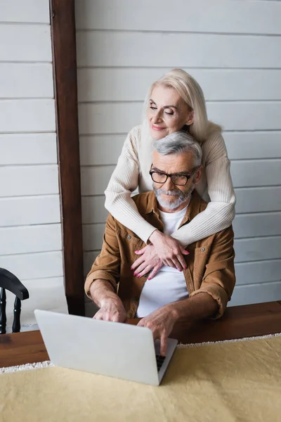 Ältere Frau umarmt Ehemann, der zu Hause am Laptop arbeitet — Stockfoto