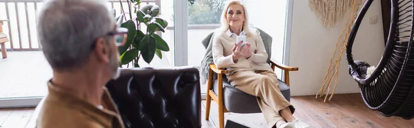 Веселая пожилая женщина, используя смартфон в кресле рядом с размытым мужем в гостиной, баннер — стоковое фото
