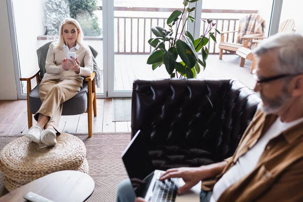 Mujer mayor usando teléfono inteligente cerca de marido borroso con portátil en el sofá - foto de stock