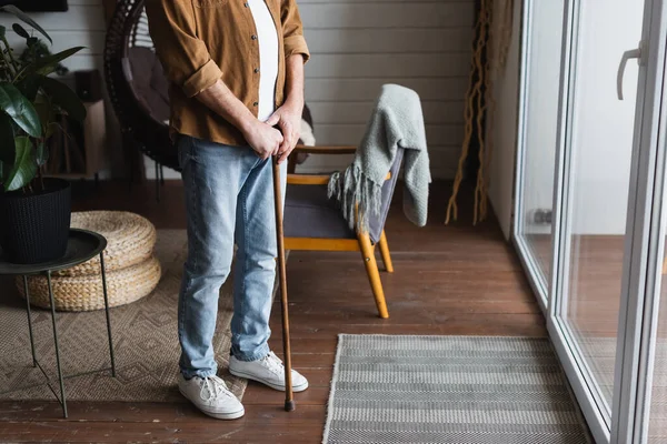 Vista cortada do homem idoso em roupas casuais segurando muleta em casa — Fotografia de Stock