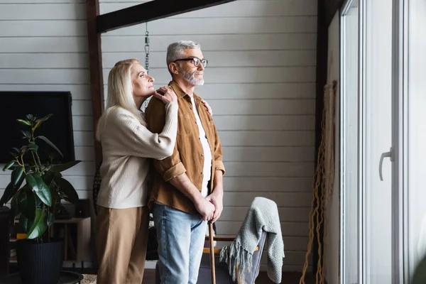 Elderly woman hugging husband with crutch at home — Stock Photo