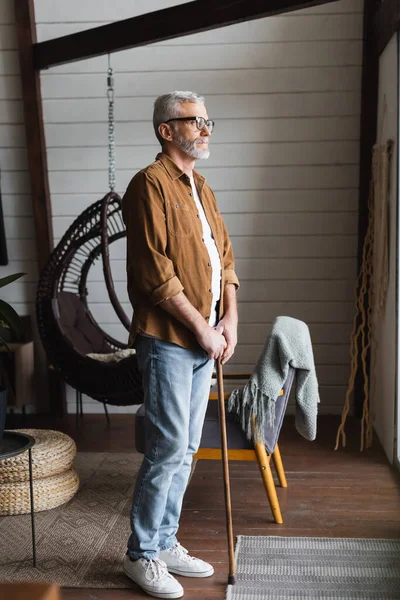 Homme âgé en lunettes tenant la béquille alors qu'il se tenait à la maison — Photo de stock