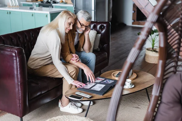 Senior couple looking at photos in album near smartphone on coffee table — Stock Photo