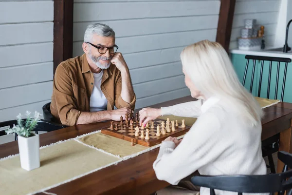 Старший мужчина играет в шахматы с размытой женой дома — стоковое фото