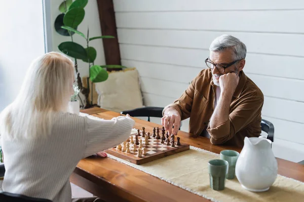 Senior couple playing chess near glasses and jug — Stock Photo