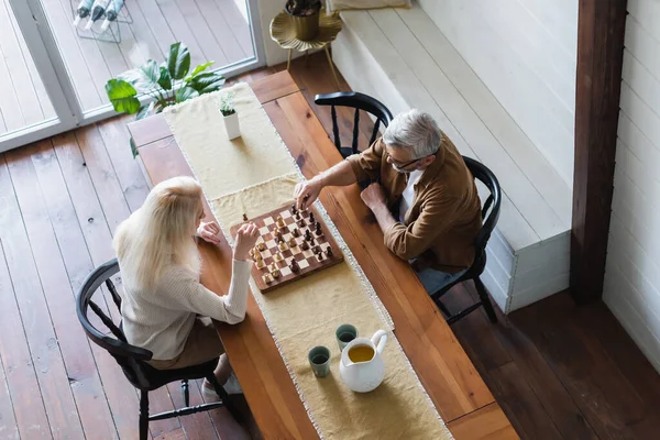 Vista aérea de pareja mayor jugando ajedrez cerca de jarra y vasos en la mesa - foto de stock