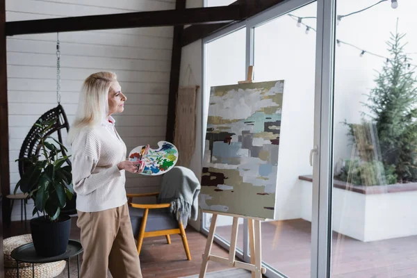 Seitenansicht einer Seniorin mit Blick auf Leinwand auf Staffelei im Wohnzimmer — Stockfoto