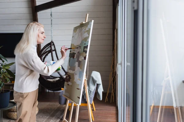 Vista lateral de la mujer mayor pintura sobre lienzo en casa - foto de stock