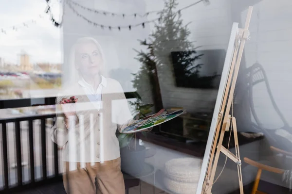 Старша жінка тримає палітру біля мольберта і вікна вдома — стокове фото