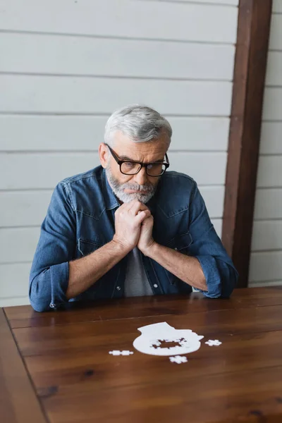 Thoughtful elderly man in eyeglasses looking at blurred puzzle on table — Stock Photo