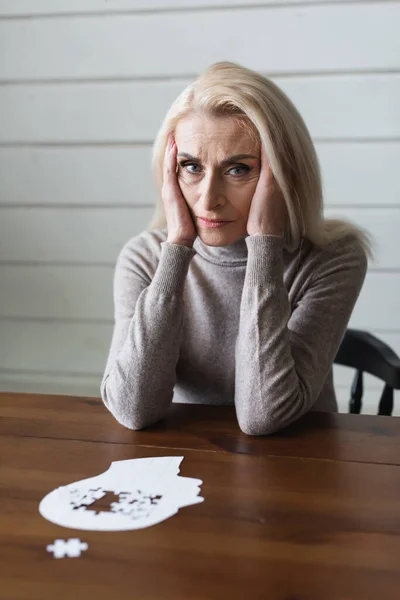 Triste femme âgée avec démence regardant la caméra près du puzzle sur le premier plan flou — Photo de stock