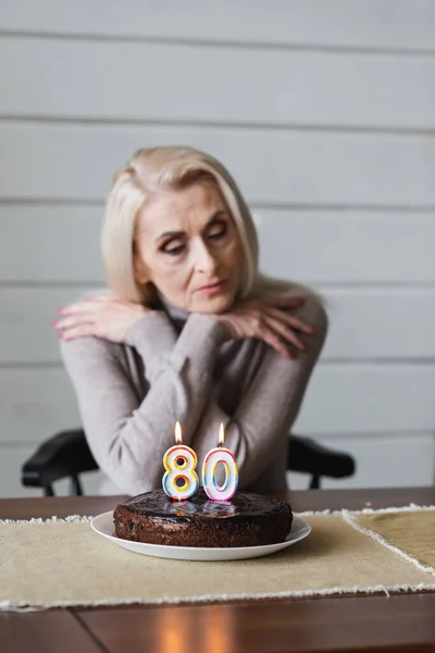 Tarta de cumpleaños con velas en forma de ochenta números en la mesa cerca de la mujer mayor borrosa - foto de stock