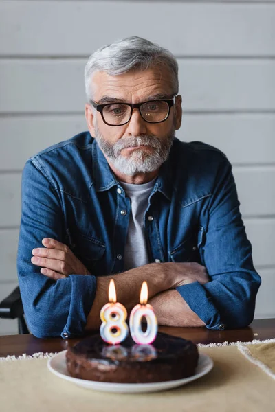 Homme seul dans des lunettes assis près du gâteau d'anniversaire flou avec des bougies en forme de quatre-vingts numéros — Photo de stock