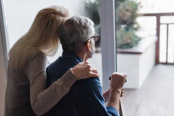 Vue arrière de la femme embrassant mari aîné avec béquille près de la fenêtre — Photo de stock