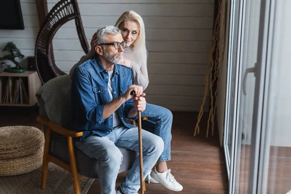 Senior man with crutch sitting on armchair near wife — Stock Photo