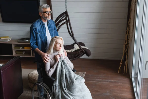 Мужчина в очках обнимает жену в инвалидном кресле дома — стоковое фото