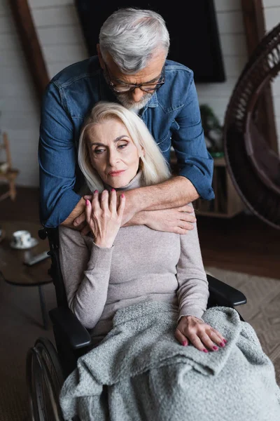 Старший мужчина в очках обнимает жену-инвалида в инвалидном кресле — стоковое фото