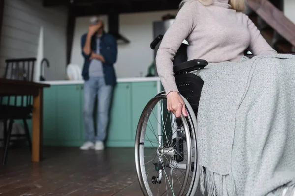 Senior woman sitting in wheelchair near husband in kitchen on blurred background — Stock Photo