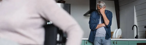 Molesto hombre mayor de pie en la cocina cerca de la esposa borrosa en casa, pancarta - foto de stock