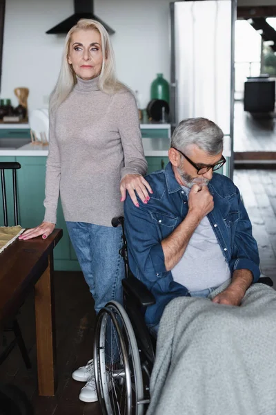 Donna anziana in piedi vicino marito scontento in sedia a rotelle con coperta — Foto stock