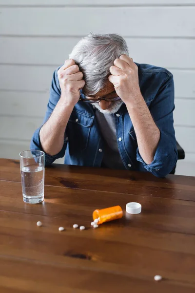 Homem deprimido olhando para pílulas borradas perto de vidro de água na mesa — Fotografia de Stock