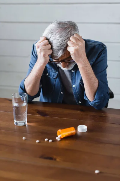 Depressed man looking at blurred pills near glass of water on table — Stock Photo