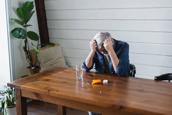 Lonely man sitting near scattered pills and water at home — Stock Photo