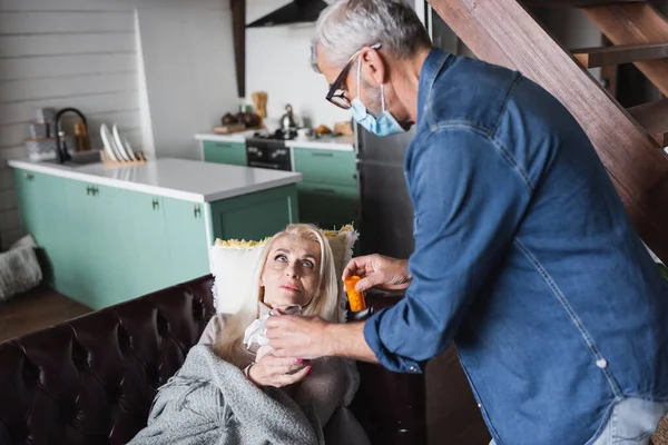 Marito offuscato in maschera medica con acqua e pillole vicino alla moglie a casa — Foto stock