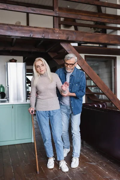 Donna anziana con stampella in piedi vicino a marito attento a casa — Foto stock