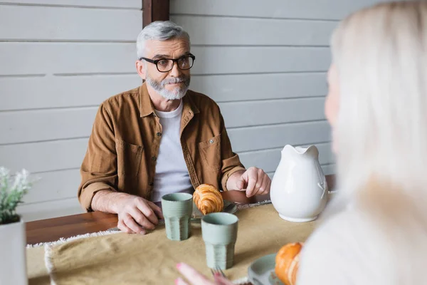 Uomo anziano guardando la moglie offuscata vicino a croissant e bicchieri sul tavolo — Foto stock