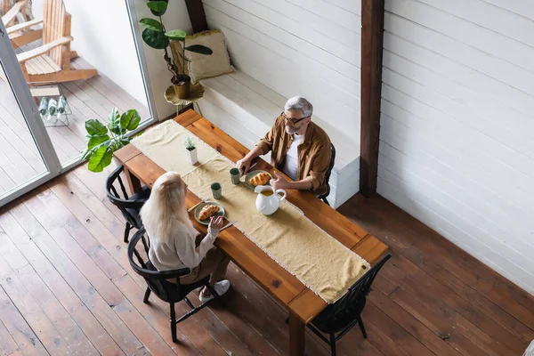 Vue aérienne du couple âgé assis près des croissants et de la cruche sur la table — Photo de stock