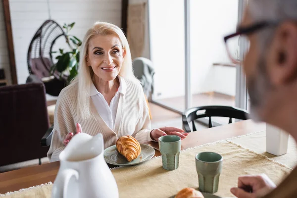 Fröhliche Seniorin schaut Ehemann bei Croissant und Gläsern an — Stockfoto