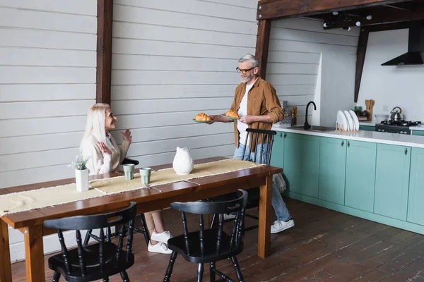 Веселая женщина сидит рядом с мужем с круассанами на кухне — стоковое фото