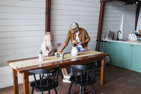 Старшая женщина смотрит на мужа с круассанами возле кувшина на столе — стоковое фото