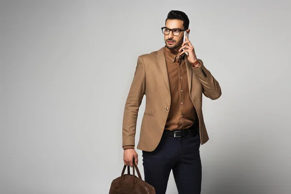 Arabischer Geschäftsmann mit Handtasche spricht auf Smartphone vor grauem Hintergrund — Stockfoto