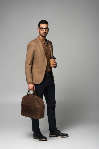 Арабский бизнесмен держит кофе, чтобы пойти и сумку на сером фоне — стоковое фото