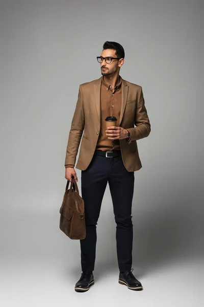 Hombre de negocios musulmán en chaqueta marrón sosteniendo taza de papel y bolso sobre fondo gris — Stock Photo