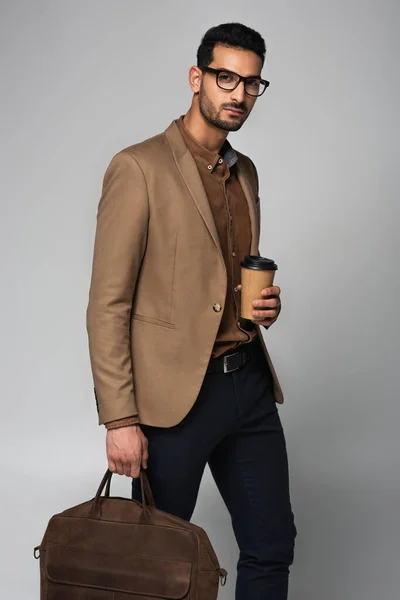 Stylish muslim businessman holding paper cup and handbag isolated on grey — Stock Photo