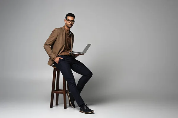Hombre de negocios musulmán con mano en el bolsillo y portátil cerca de la silla sobre fondo gris - foto de stock