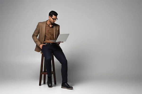 Арабский бизнесмен смотрит на ноутбук и сидит на стуле на сером фоне — стоковое фото