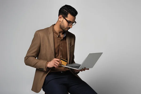 Homem muçulmano em uso formal segurando laptop e cartão de crédito isolado em cinza — Fotografia de Stock