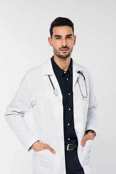 Médico árabe con estetoscopio cogido de la mano en bolsillos de abrigo blanco aislado en gris - foto de stock