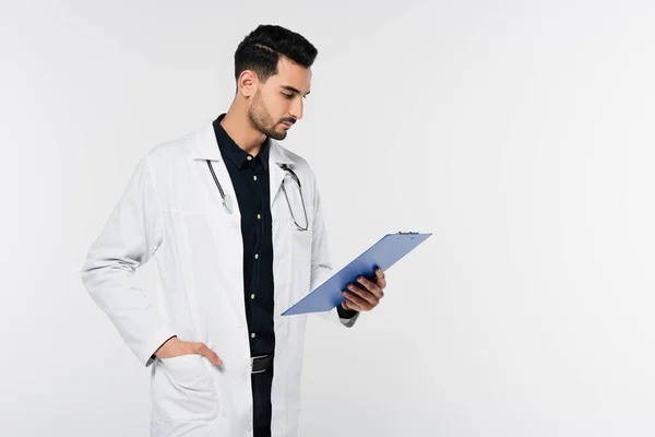 Médico árabe mirando el portapapeles aislado en gris - foto de stock