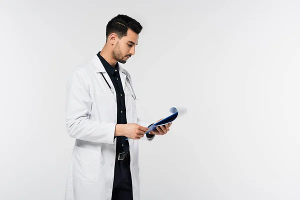 Вид збоку арабського лікаря, який дивиться на буфер обміну з паперами ізольовано на сірому — стокове фото