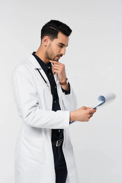 Vista lateral del médico musulmán pensativo que mira el diagnóstico en el portapapeles aislado en gris - foto de stock