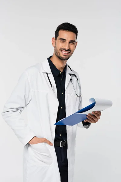 Cheerful arabian doctor holding clipboard and looking at camera isolated on grey — Stock Photo
